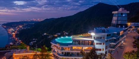 Hotel de vários andares com piscina no terraço, rodeado por montanhas e uma paisagem urbana costeira.