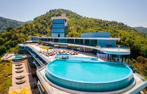 Un hotel moderno con una gran piscina central, mobiliario exterior y un paisaje montañoso de fondo.