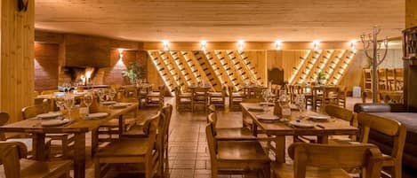 A restaurant with wooden tables and chairs, a brick wall with a fireplace, and a sloped ceiling with wooden beams.