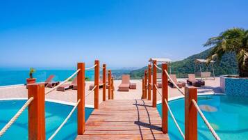 A wooden deck leading to a pool with lounge chairs and umbrellas, overlooking a coastal view.