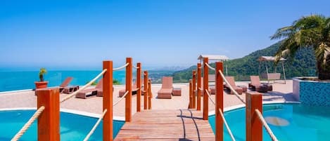 A wooden deck leading to a pool with lounge chairs and umbrellas, overlooking a coastal view.