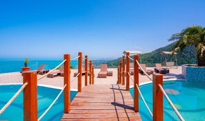 A wooden deck leading to a pool with lounge chairs and umbrellas, overlooking a coastal view.