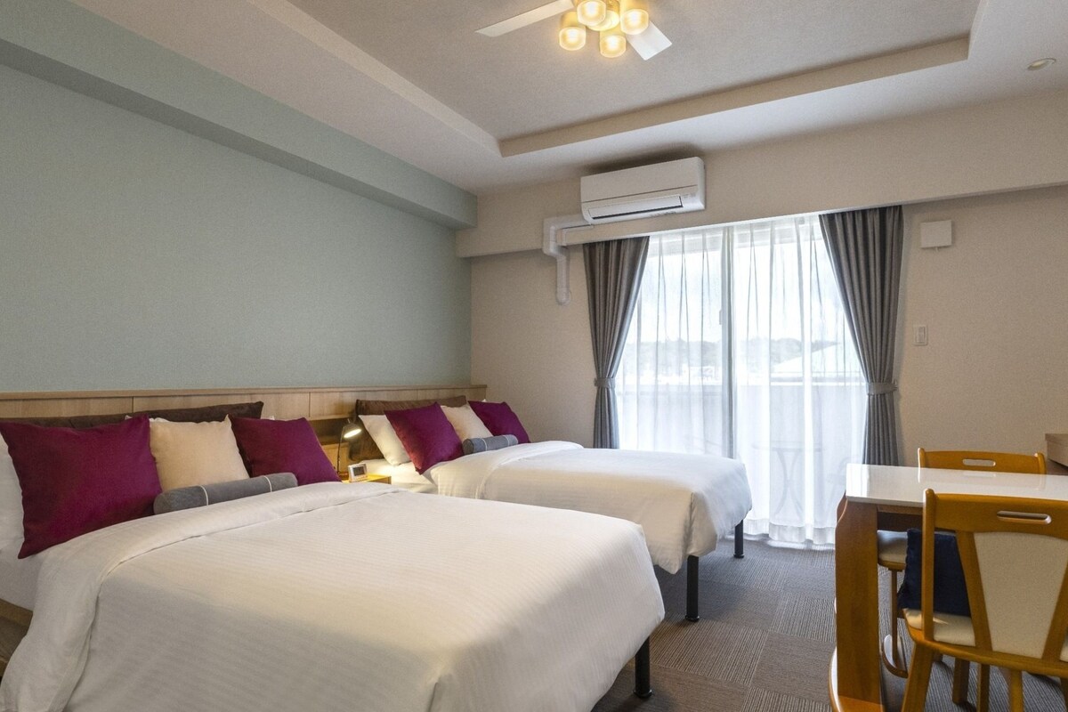 Une chambre d’hôtel avec deux lits, un bureau, une chaise, un appareil de climatisation et une fenêtre avec des rideaux.