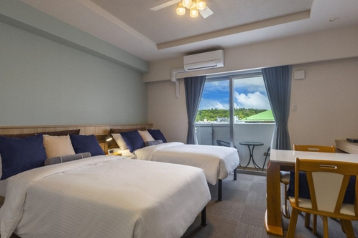 A hotel room with two beds, a balcony with a table and chairs, and an air conditioning unit.