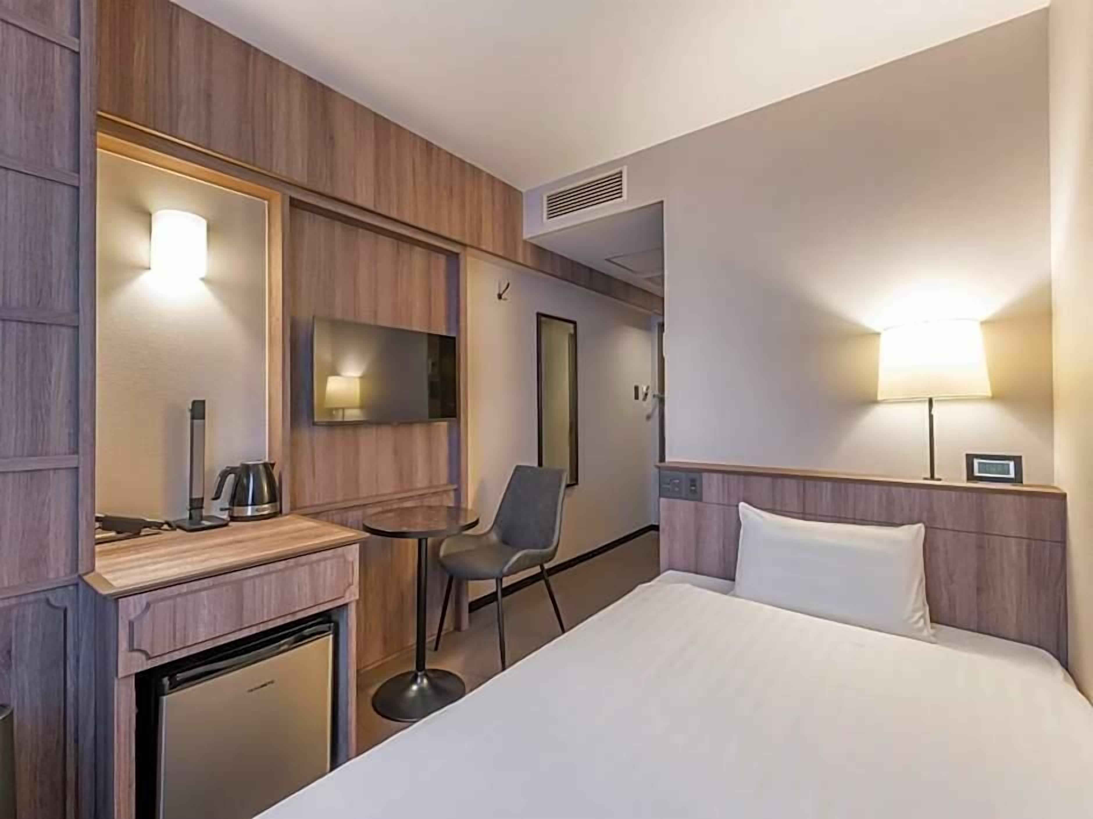 A hotel room with a bed, a desk with a kettle, a chair, a TV, and a lamp.