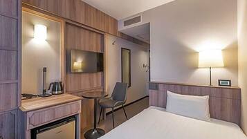A hotel room with a bed, a desk with a kettle, a chair, a TV, and a lamp.