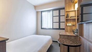 A hotel room with a bed, a small table, a chair, a desk, a shelf with items, and a window with curtains.