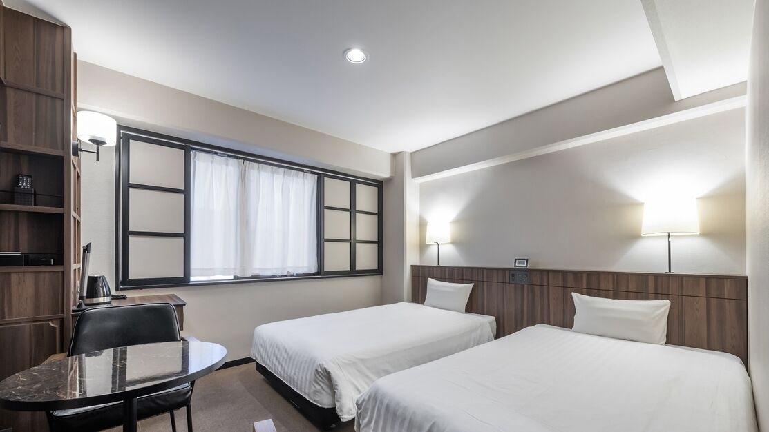 Habitación de hotel con dos camas, un escritorio con silla, una ventana con cortinas y lámparas fijadas a la pared.