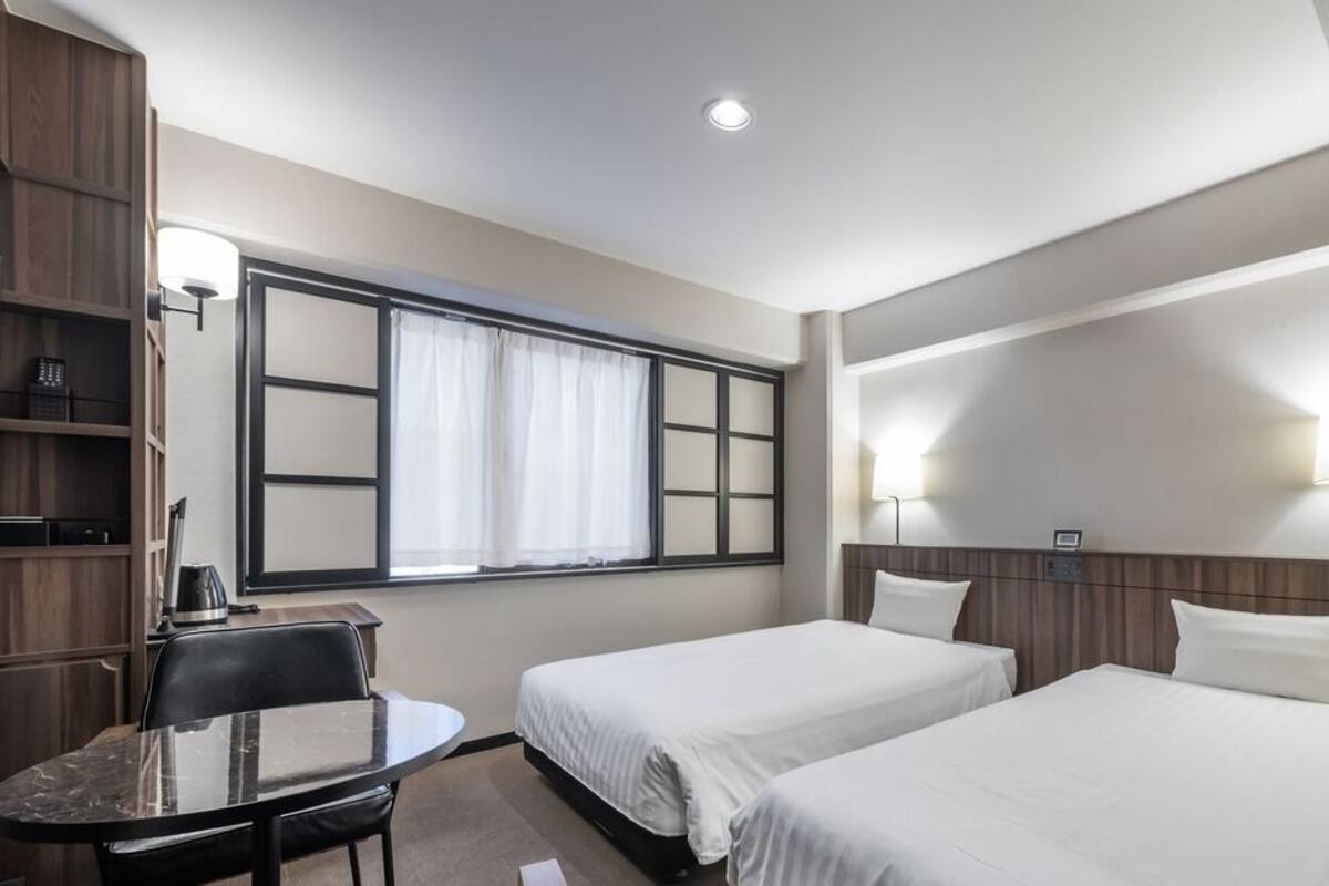Une chambre d’hôtel avec deux lits, un bureau avec une chaise, une fenêtre avec des rideaux et des lampes fixées au mur.