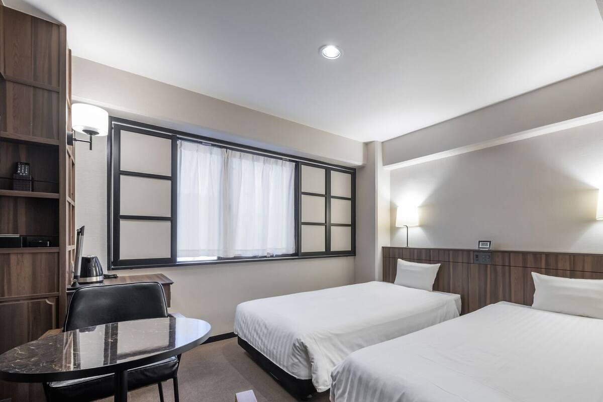 Une chambre d’hôtel avec deux lits, un bureau, une chaise, une fenêtre avec des rideaux et des lampes fixées au mur.