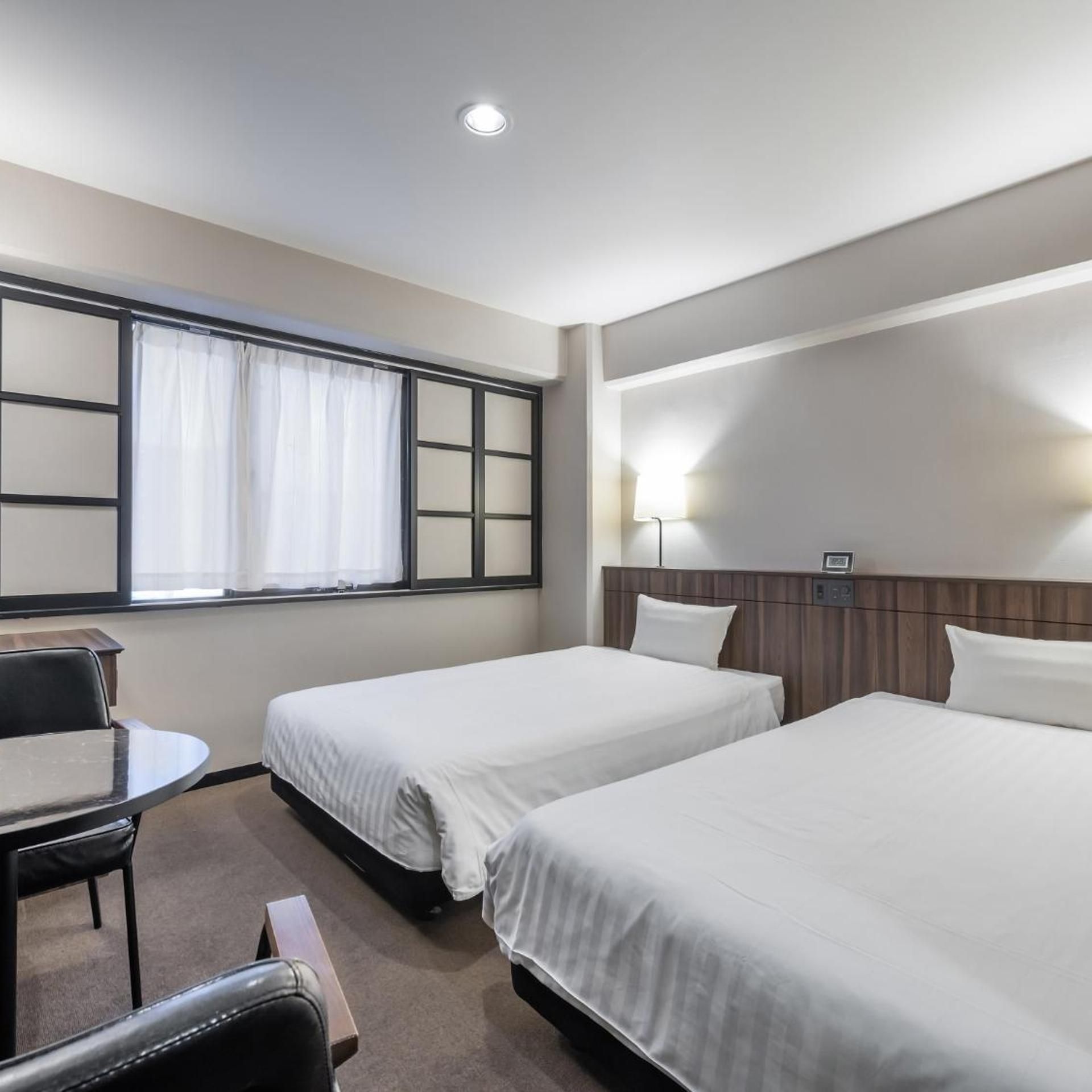 A hotel room with two single beds, a desk, a chair, and a window with curtains.