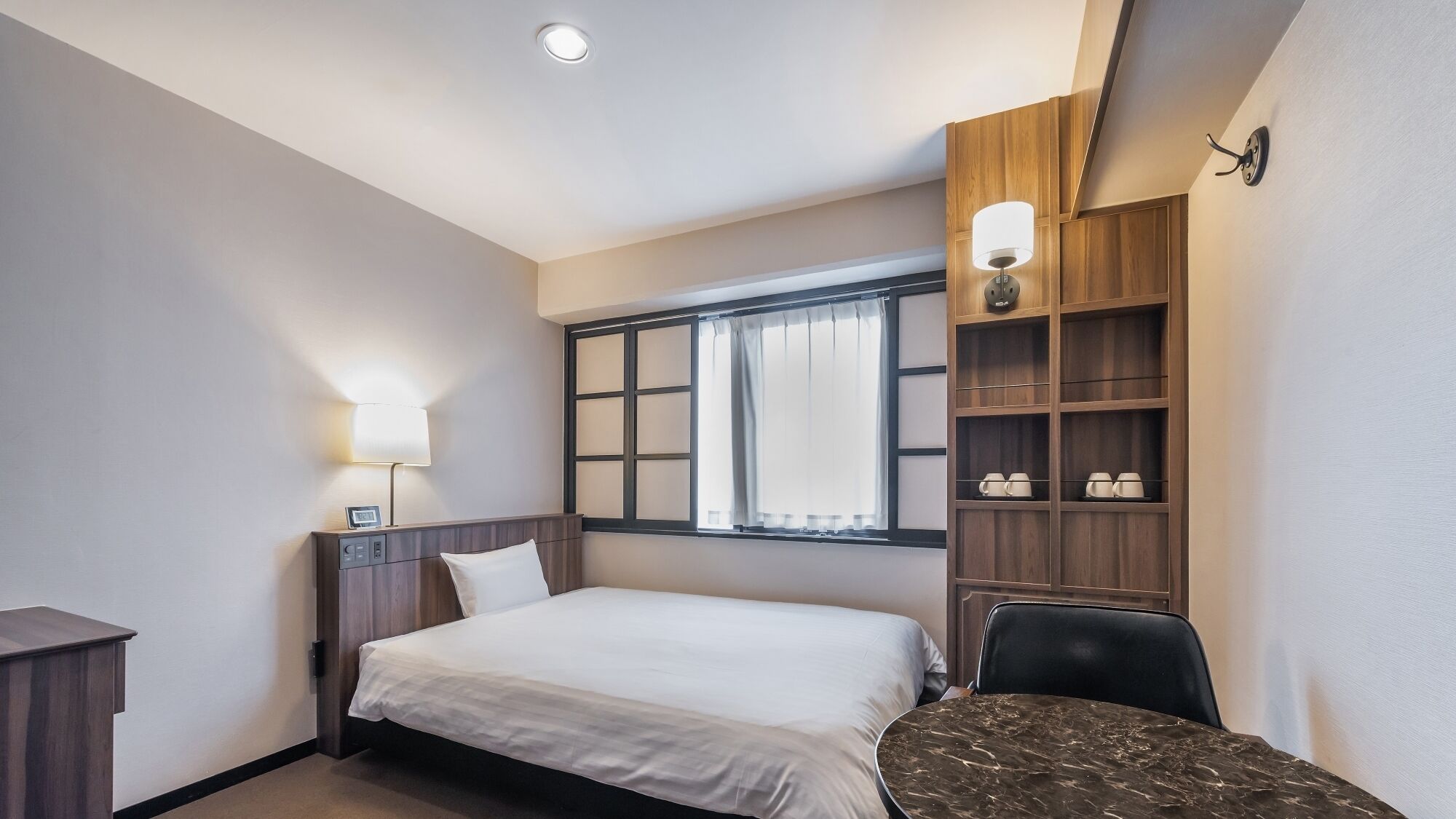Habitación de hotel con cama, mesita de noche, lámpara, ventana con cortinas y estante con tazas.