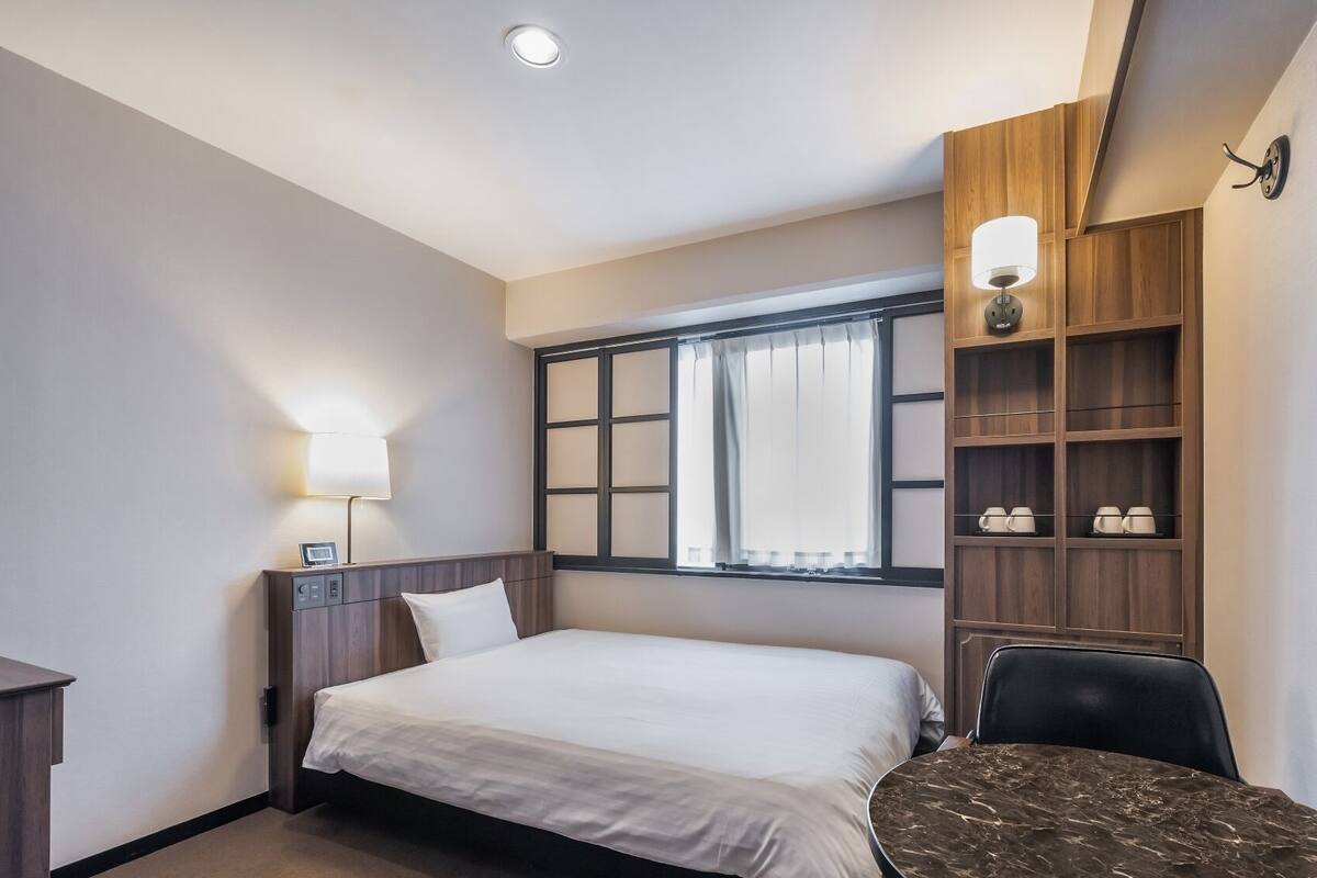 Une chambre d’hôtel comprenant un lit, une table de chevet, une lampe, une fenêtre avec des rideaux et une étagère avec des tasses.