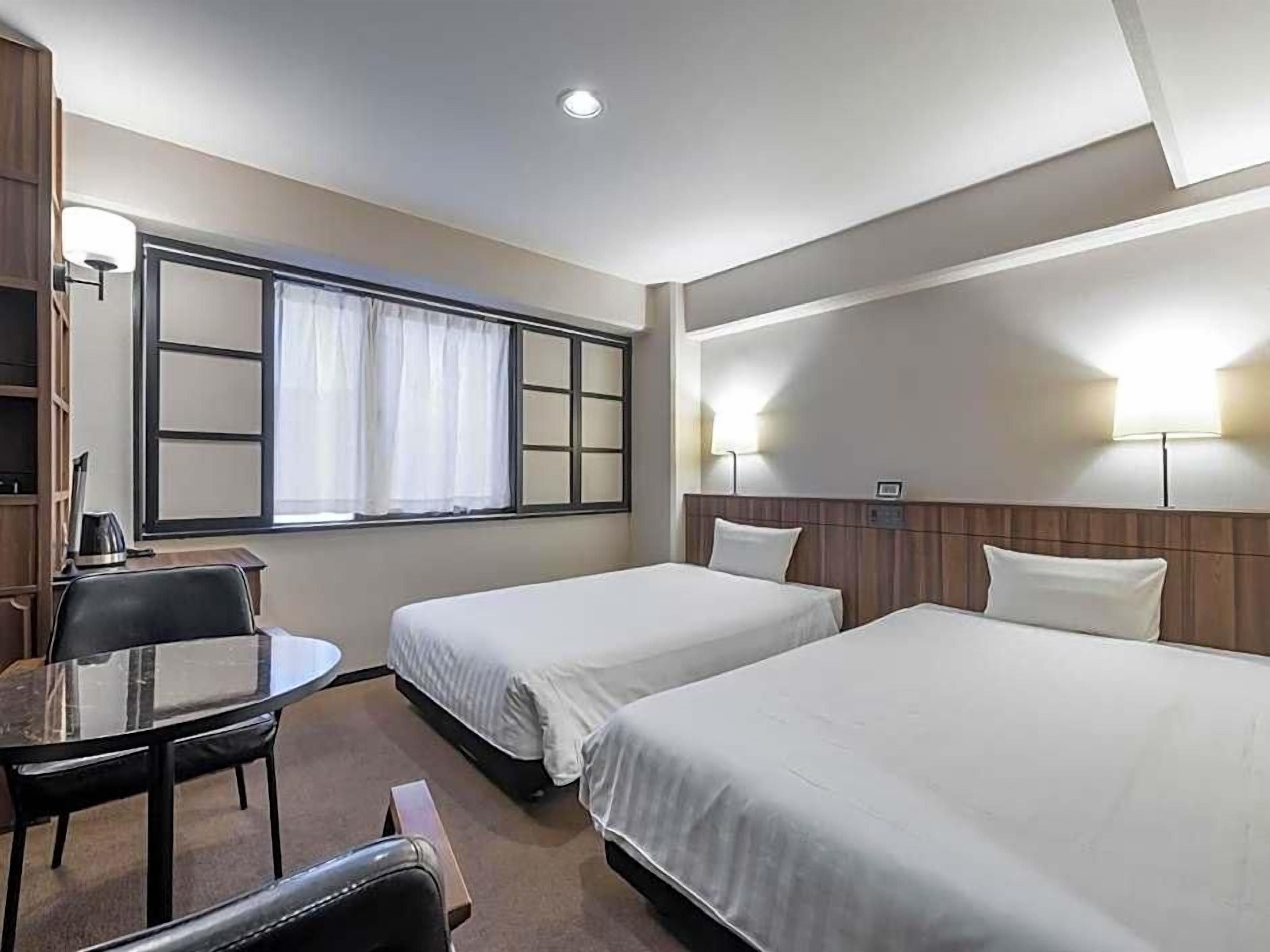 A hotel room with two single beds, a small desk with a chair, a window with curtains, and a wall-mounted lamp.