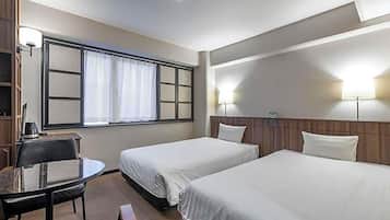 A hotel room with two single beds, a small desk with a chair, a window with curtains, and a wall-mounted lamp.