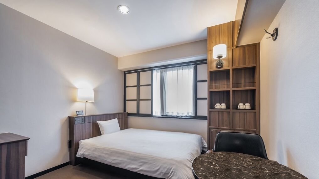 Habitación de hotel con cama, mesita de noche, lámpara, ventana con cortinas, estanterías empotradas y un escritorio con silla.