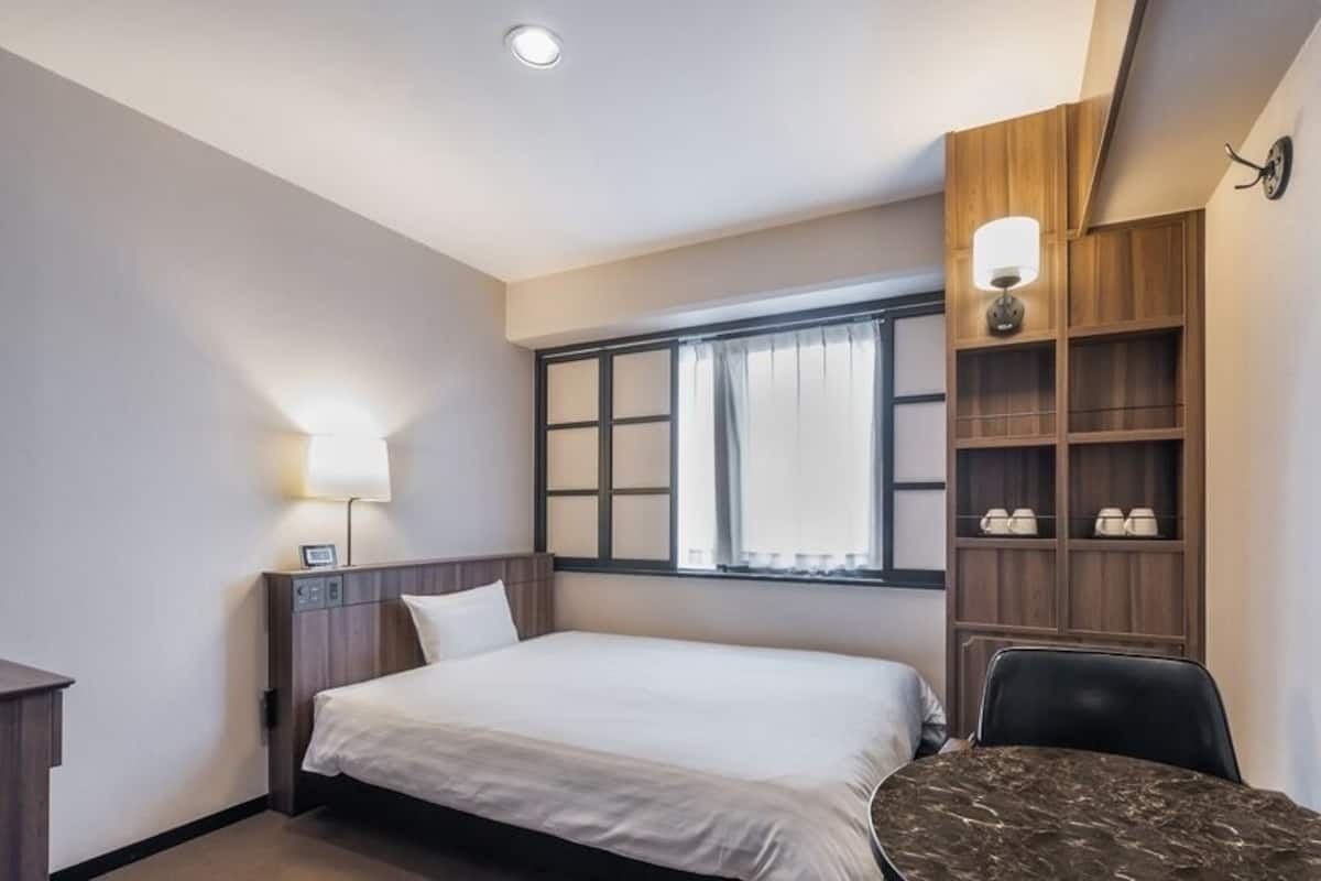 Habitación de hotel con cama, mesita de noche, lámpara, ventana con cortinas, estanterías empotradas y un escritorio con silla.