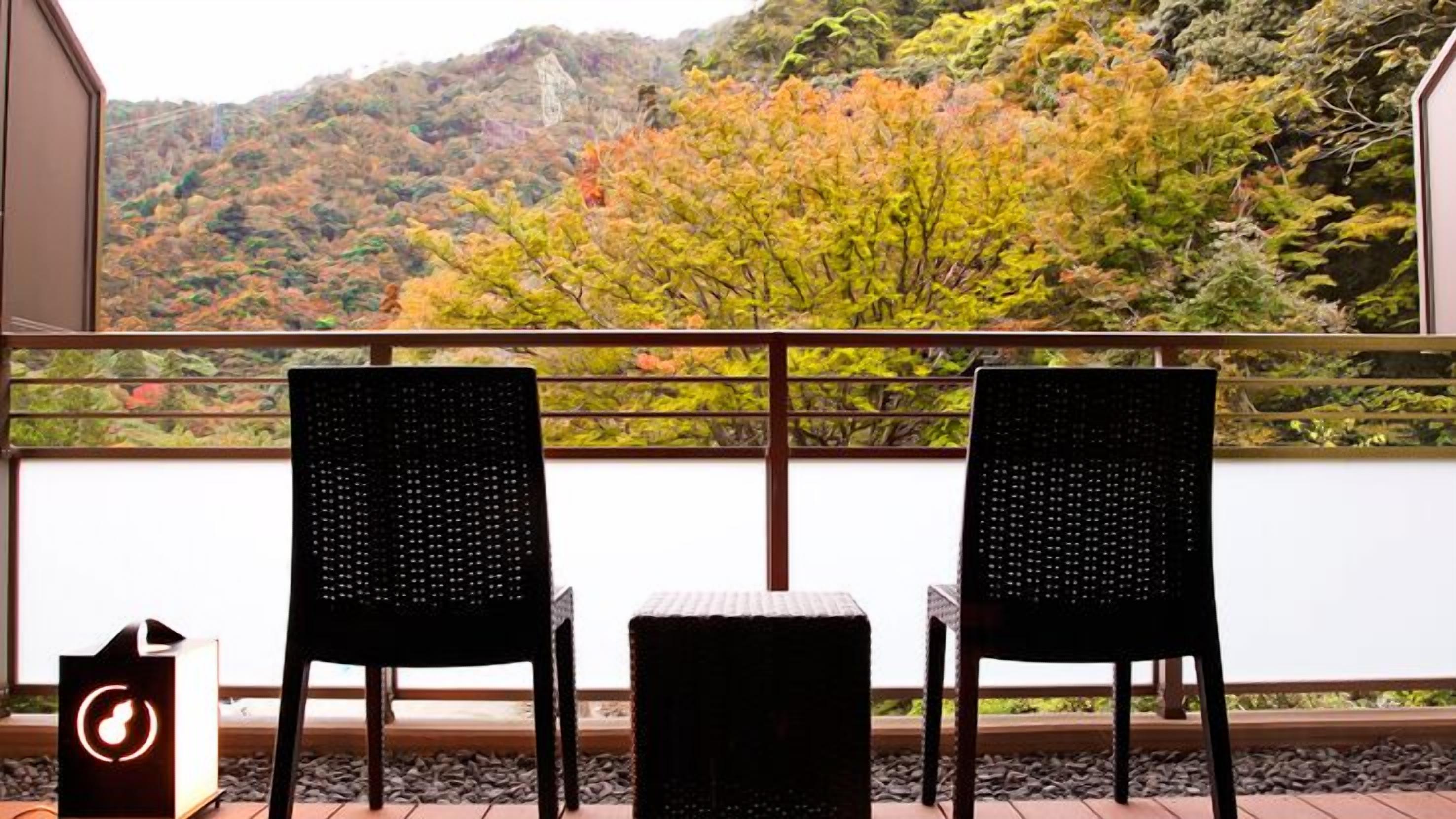 A balcony with two wicker chairs and a small table, overlooking a mountainous landscape with autumn foliage.