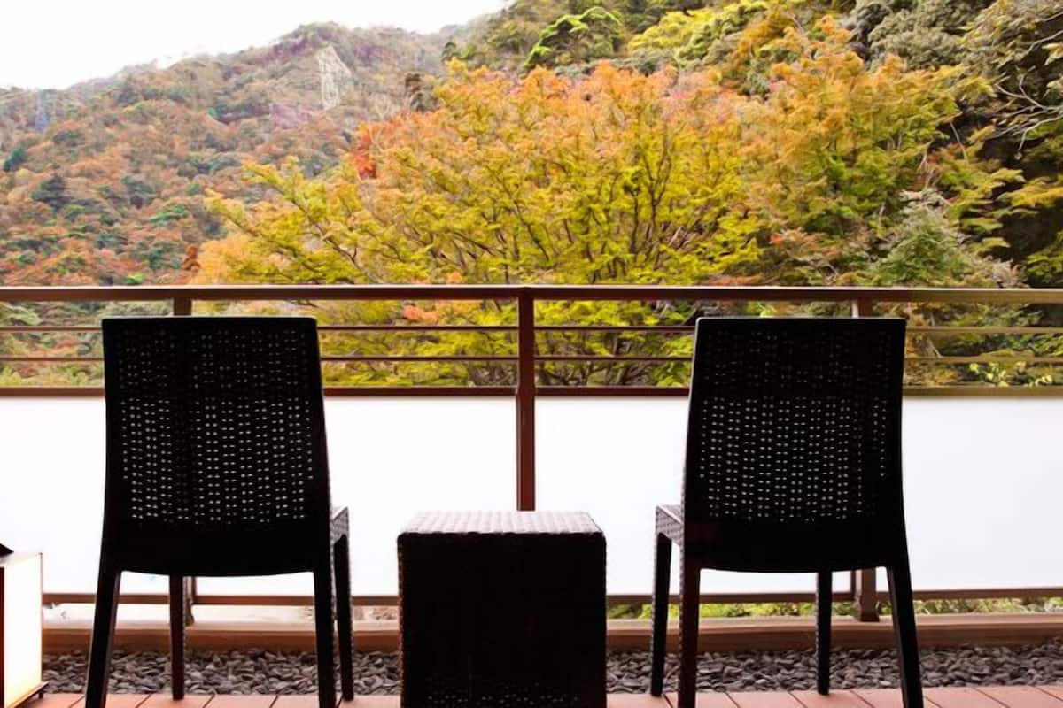 A balcony with two wicker chairs and a small table, overlooking a mountainous landscape with autumn foliage.