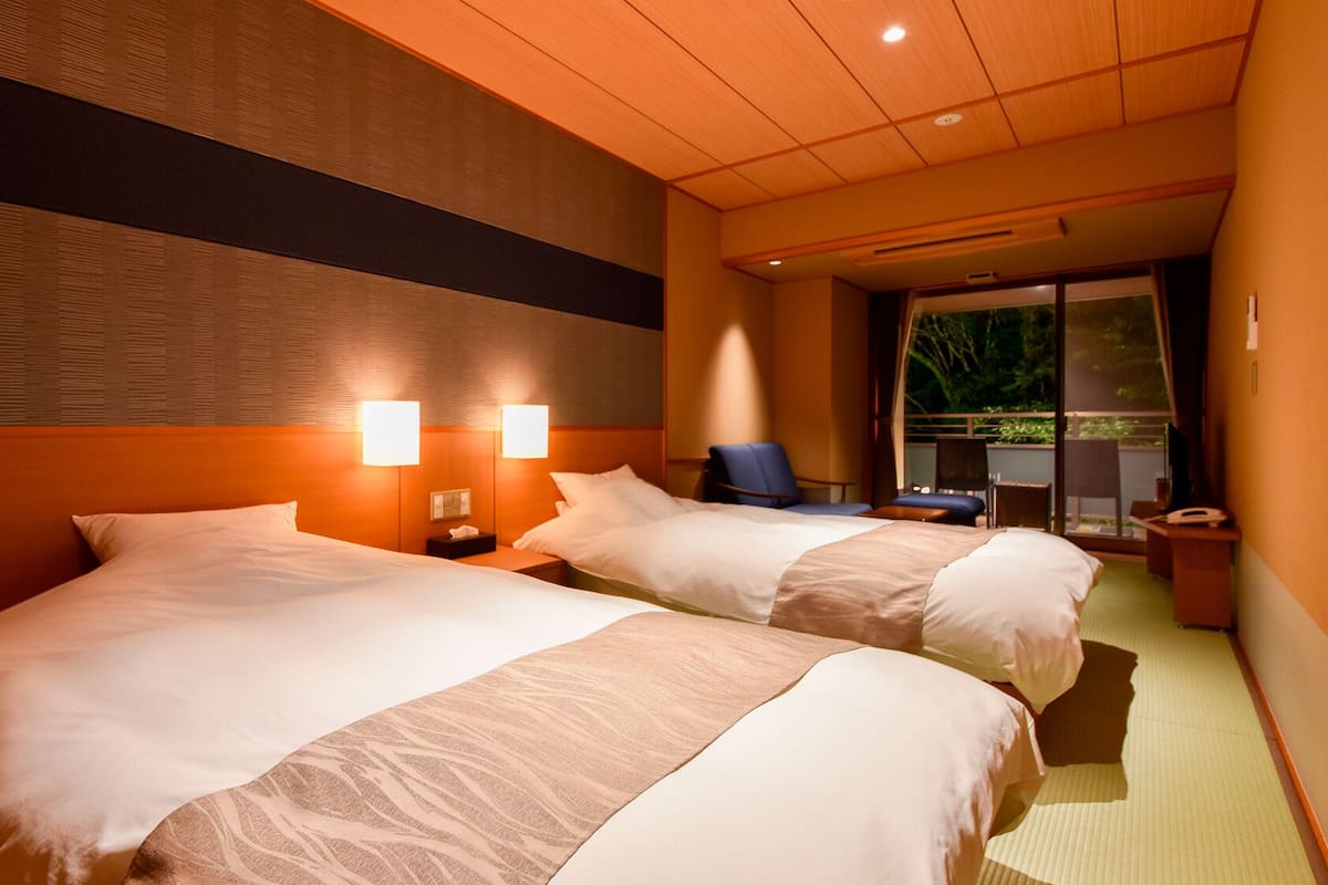 A hotel room with two beds, a desk, a chair, and a view of greenery outside.