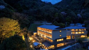 Un edificio de varios pisos con techo plano, rodeado de un bosque frondoso, con varios balcones e iluminación exterior.