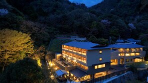 Un edificio de varios pisos con techo plano, rodeado de un bosque frondoso, con varios balcones e iluminación exterior.