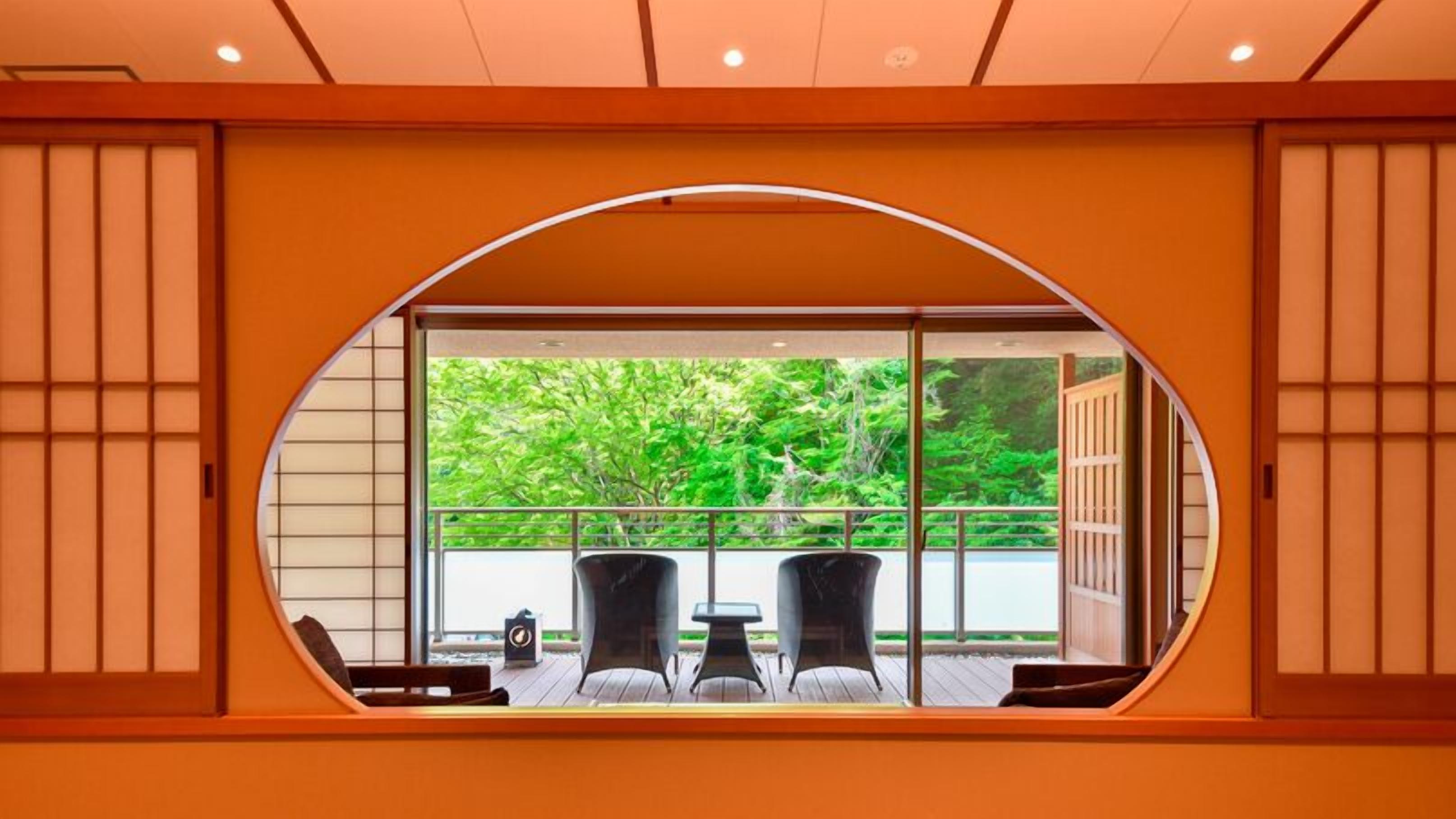 Una habitación con un ventanal ovalado grande, dos sillas, una mesita y vistas al exterior con vegetación.