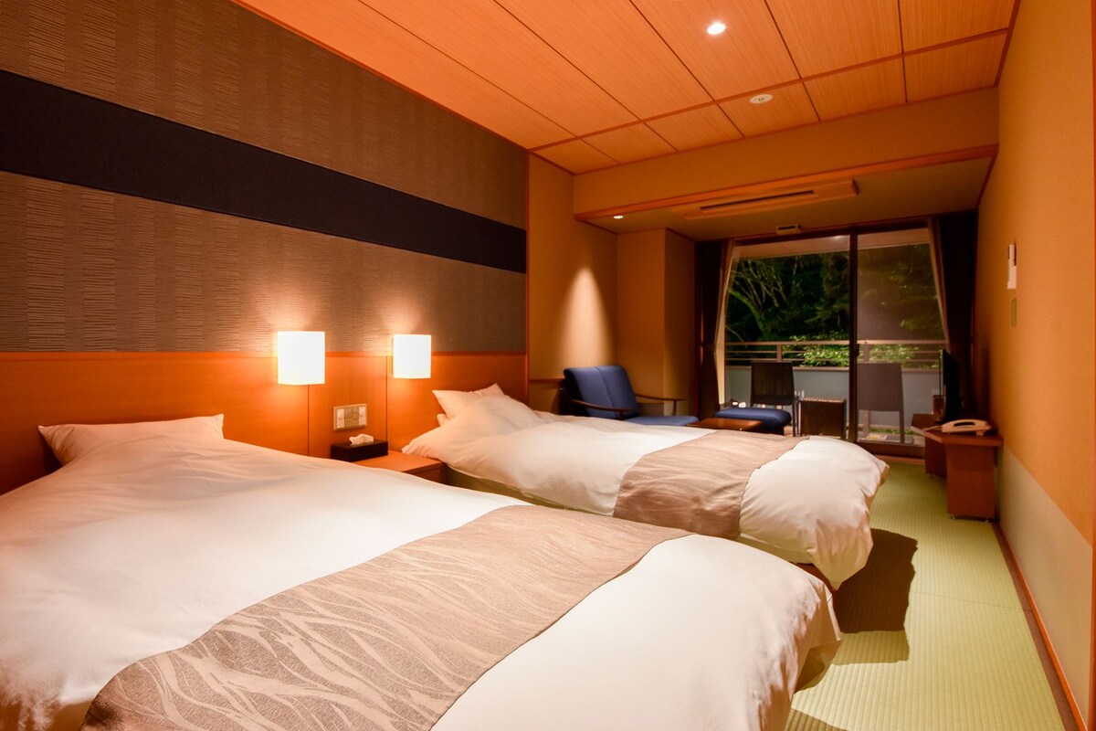Quarto de hotel com duas camas, uma escrivaninha, uma cadeira e vista para o verde lá fora.