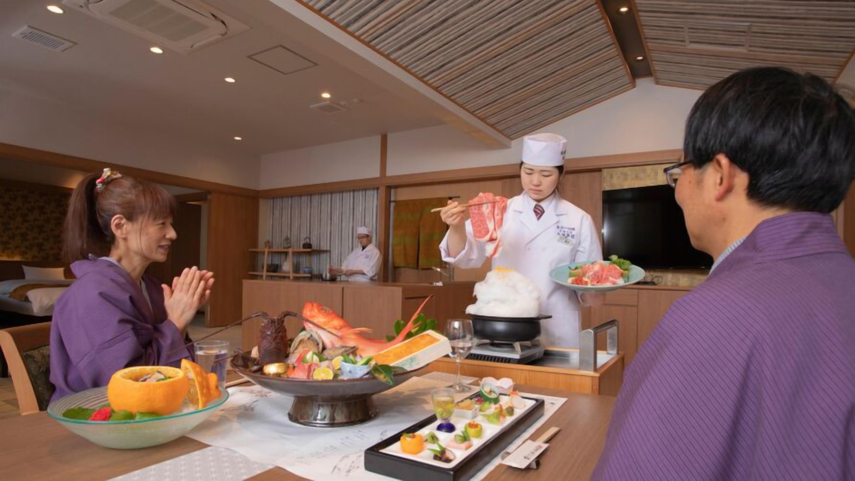 Un comedor con un chef sirviendo comida, una mesa de comedor con diversos platos y una cocina al fondo.