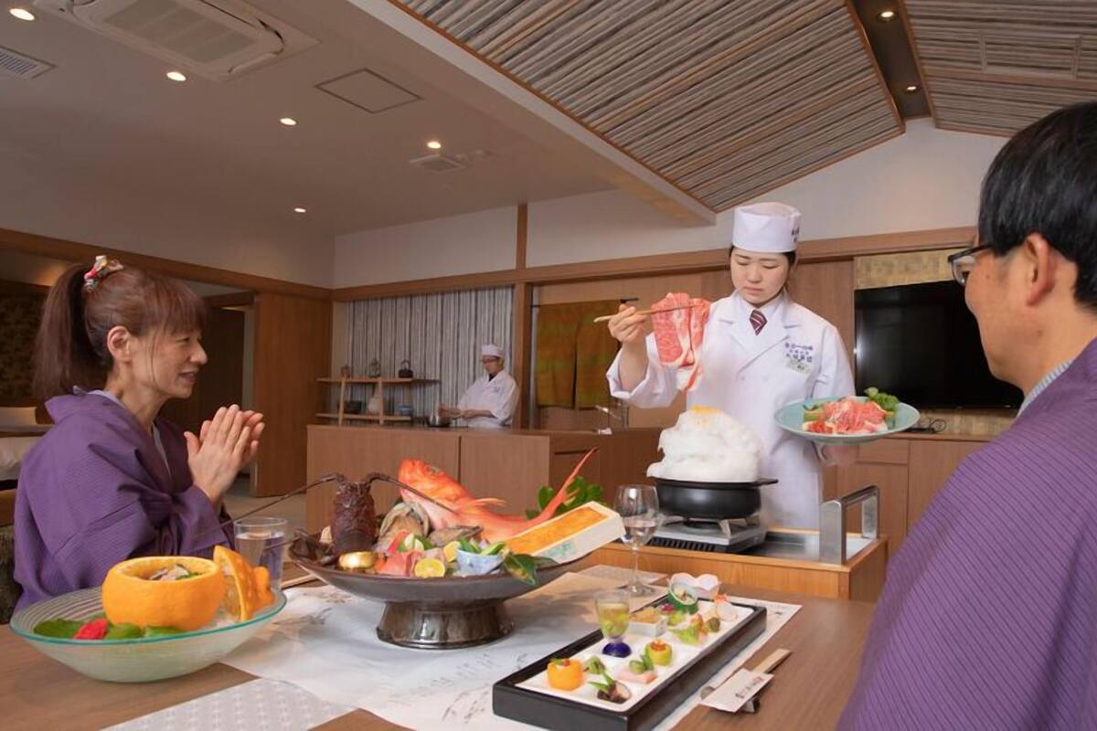 A dining area with a chef serving food, a dining table with various dishes, and a kitchen in the background.