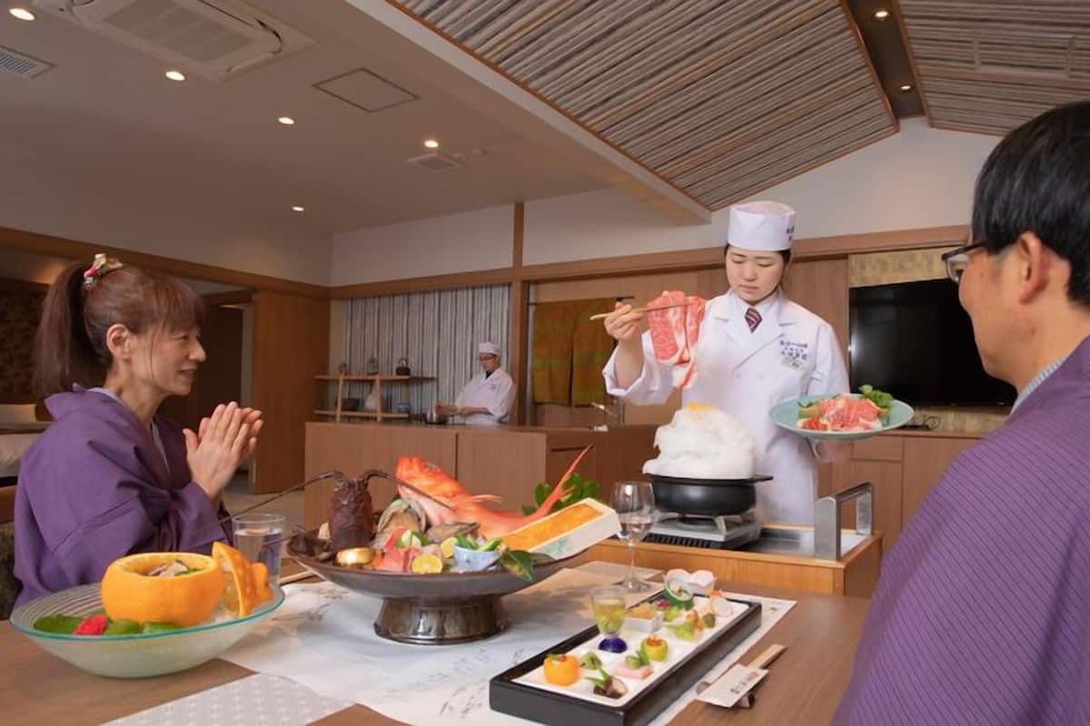 A dining area with a chef serving food, a dining table with various dishes, and a kitchen in the background.