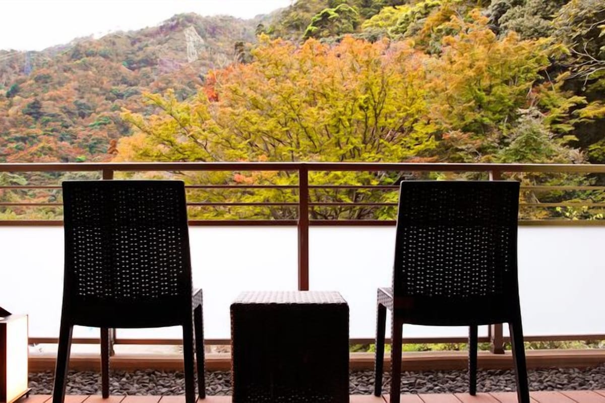 A balcony with two wicker chairs and a small table, overlooking a mountainous landscape with autumn foliage.