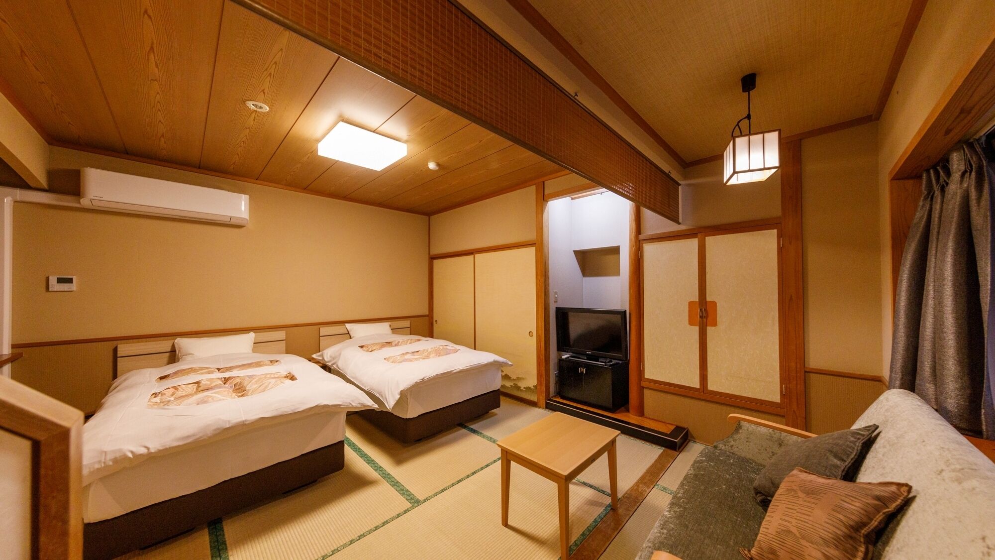 A traditional Japanese-style room with two beds, a television, a sofa, tatami flooring, and a sliding door.