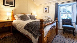 A neatly made bed with a wooden headboard, a bedside table with a lamp, a chair, and a window with curtains. - The Strands & Screes Inn (Seascale)