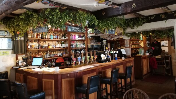 A bar with wooden bar stools, shelves of bottles, and a counter with a computer and a cash register. - The Strands & Screes Inn (Seascale)