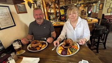 Two people seated at a wooden table with plates of food and a glass of beer. The setting appears to be a bar or pub with shelves of bottles in the background.
