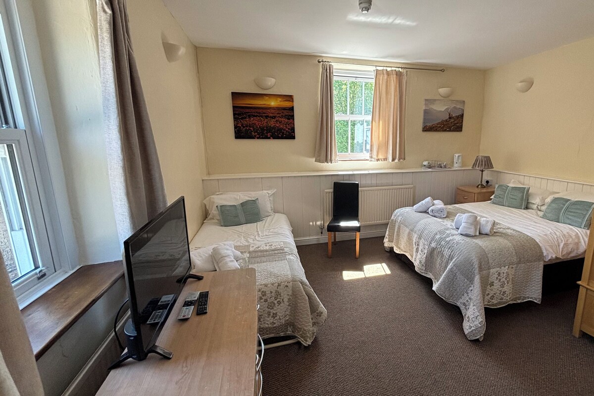 Une chambre avec deux lits, un bureau avec un ordinateur et des télécommandes, une chaise, un radiateur et une fenêtre avec des rideaux.