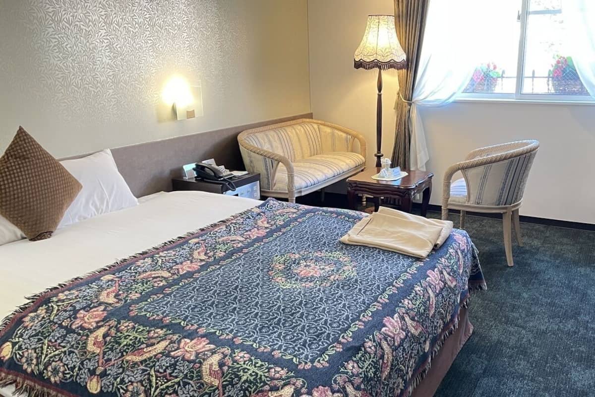 Ein Hotelzimmer mit Bett, Stuhl, Nachttisch, Lampe und einem Fenster mit Vorhängen.