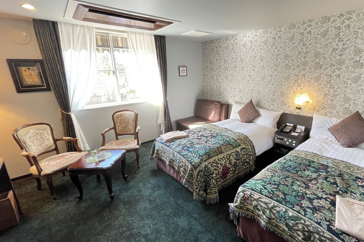 A hotel room with two beds, a desk, a chair, and a window with curtains.