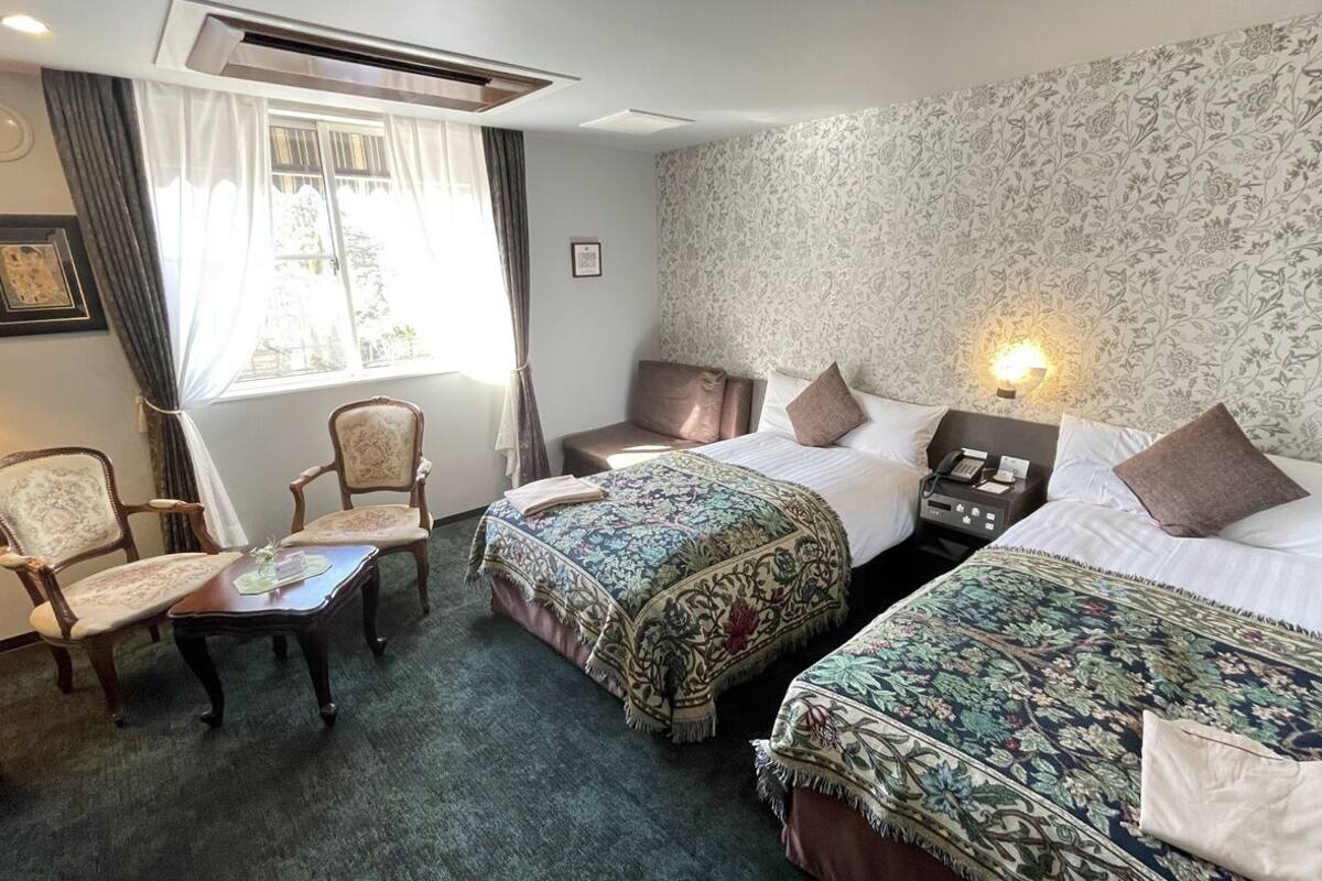 Ein Hotelzimmer mit zwei Betten, einem Sessel, einem kleinen Tisch und einem Fenster mit Vorhängen.