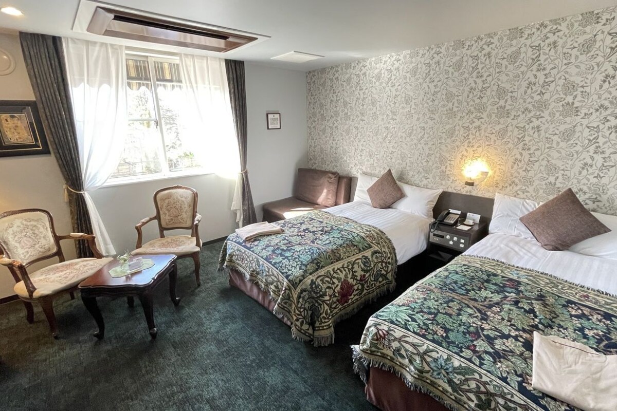 A hotel room with two beds, a chair, a small table, and a window with curtains.