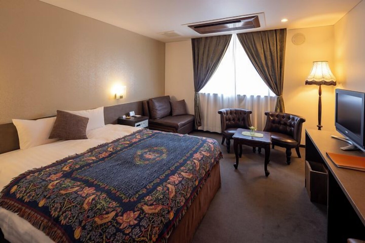 A hotel room with a large bed, a desk, a chair, a TV, and a window with curtains.