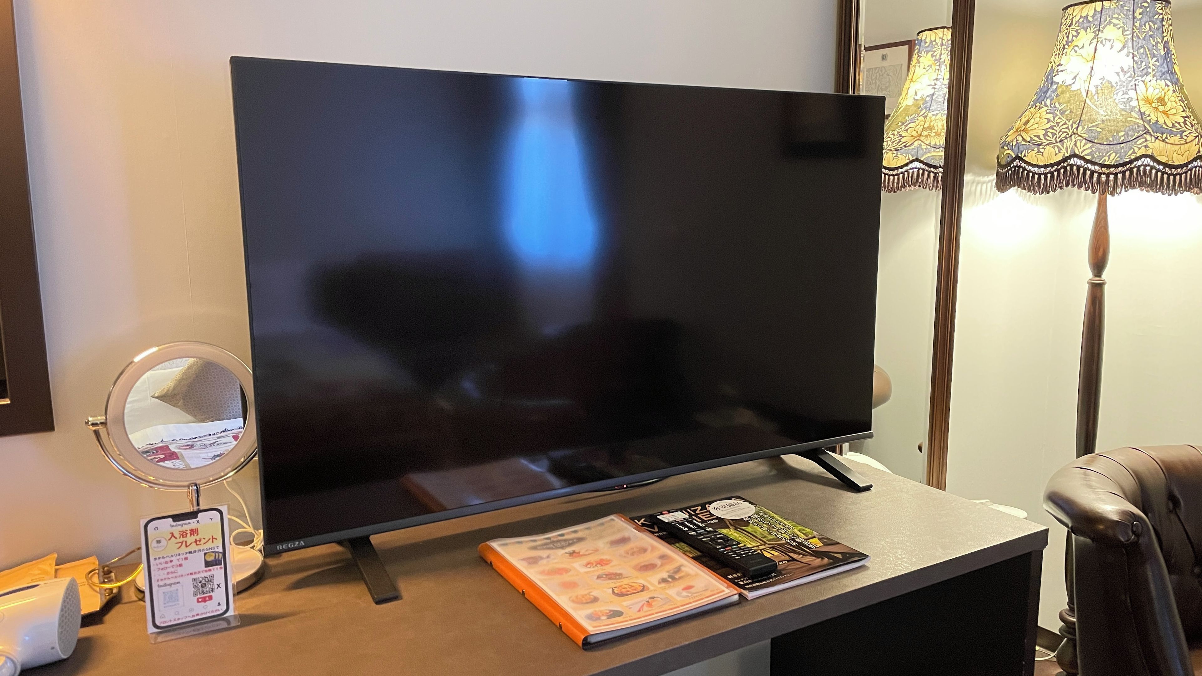 Un televisor de pantalla plana sobre un soporte, una lámpara, un espejo y una revista sobre una mesa.