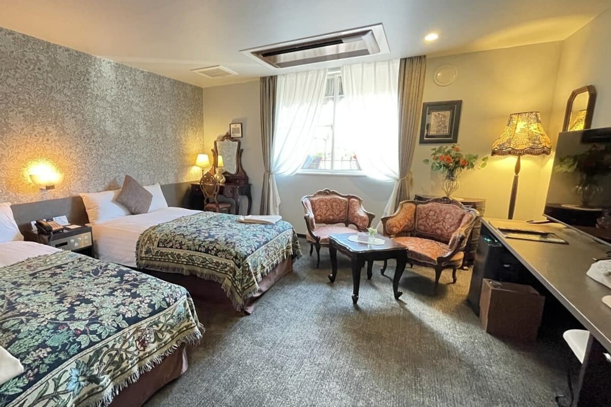 Ein Hotelzimmer mit zwei Betten, einem Schreibtisch, einem Stuhl, einem Tisch und einem fenster mit Vorhängen.