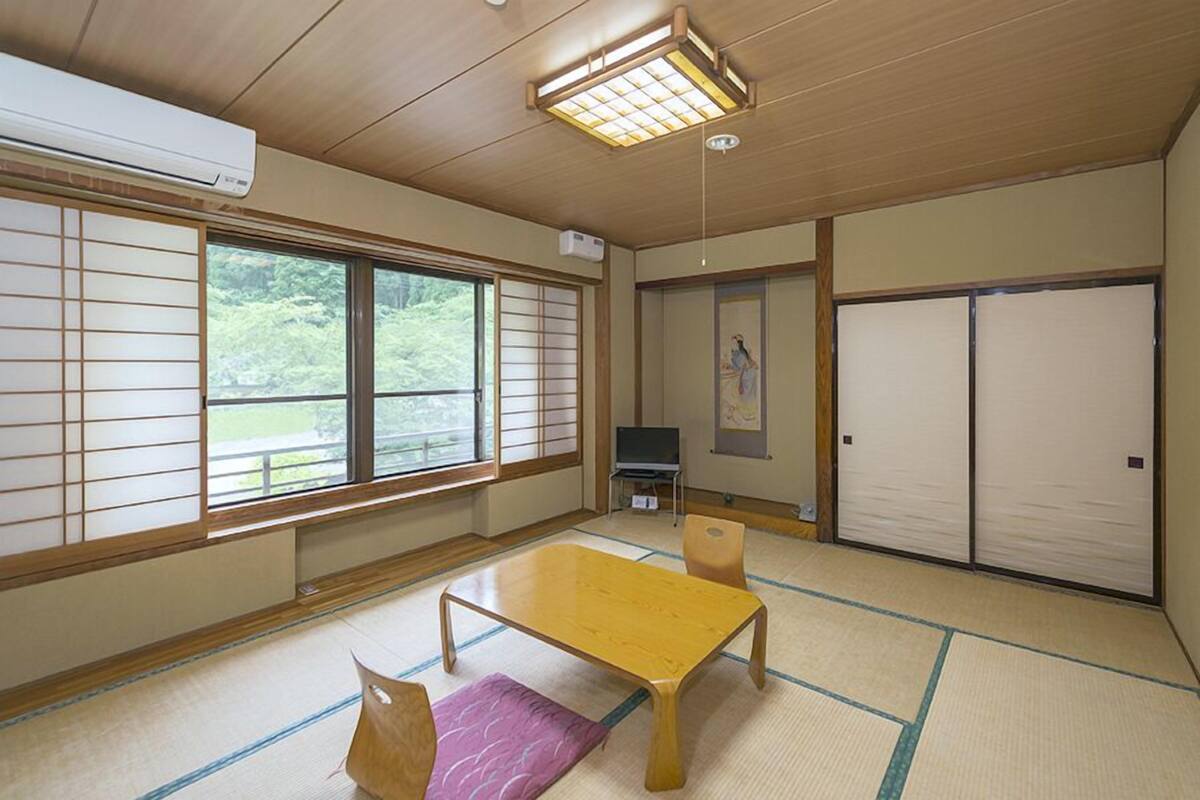 Een traditionele Japanse kamer met tatami vloeren, een houten tafel, een televisie, schuifdeuren en uitzicht op groen buiten.