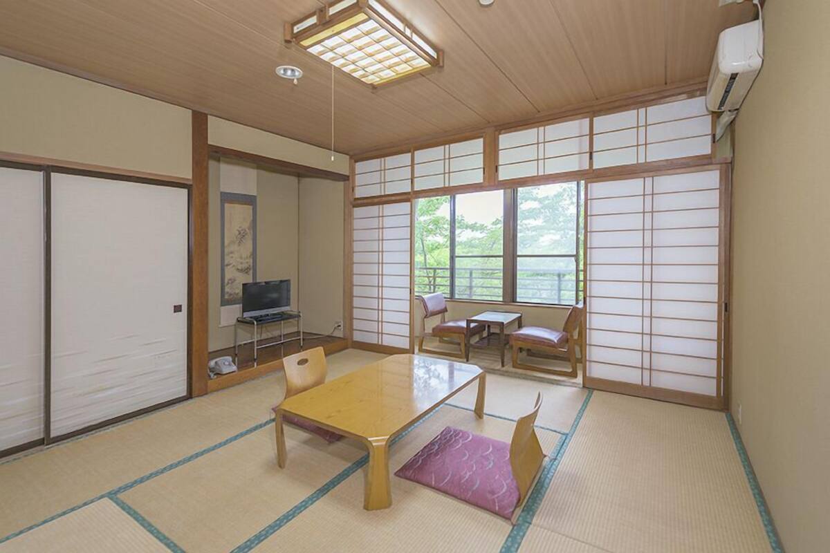 Een traditionele Japanse kamer met tatami vloerbedekking, een lage tafel, een televisie, schuifdeuren en uitzicht op groen.