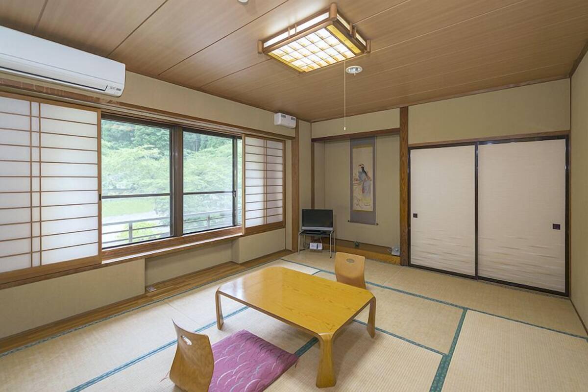 Een traditionele Japanse kamer met tatami vloeren, een lage tafel, een televisie, schuifdeuren en uitzicht op groen buiten.