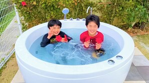 Two individuals in a hot tub with a metal fence and greenery in the background. - Villa Charme (Motobu)