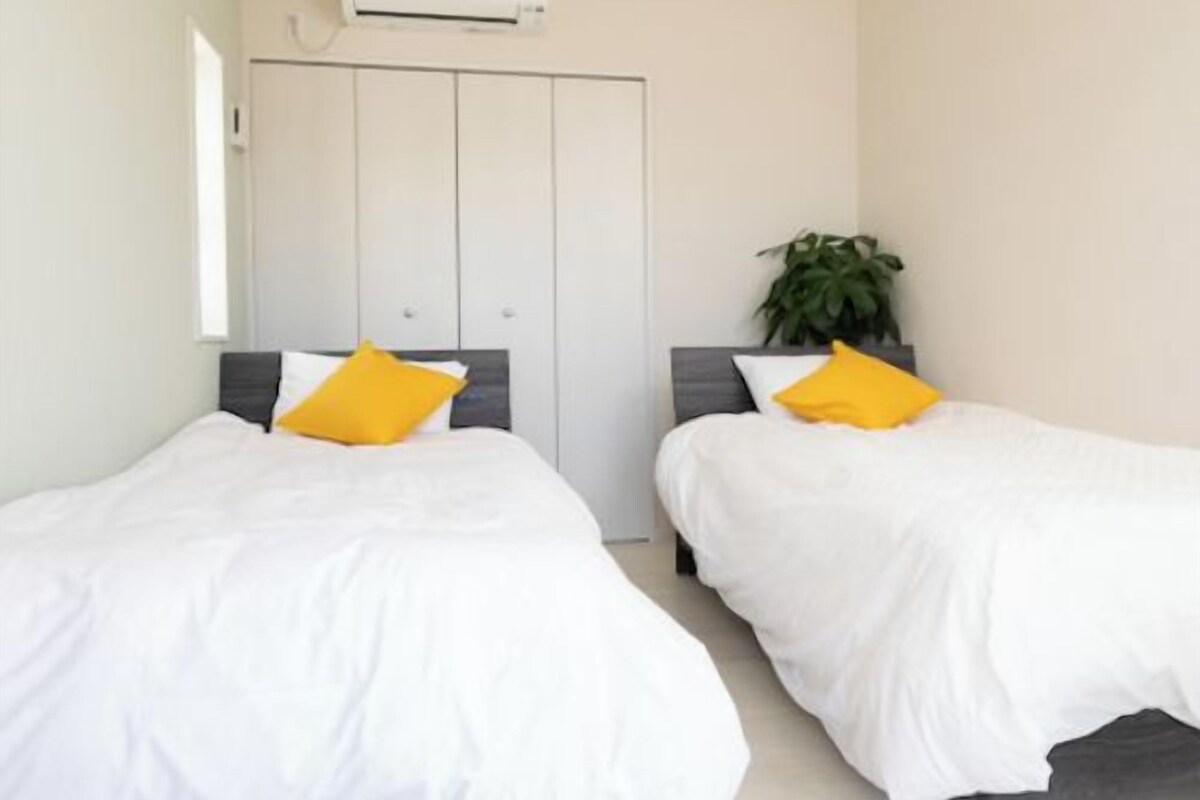 A bedroom with two beds, each with white bedding and a single yellow pillow. There is a potted plant on a table beside one bed.