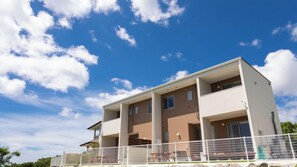 A two-story building with a balcony on the second floor, a covered outdoor seating area, and a white fence in the foreground. - Villa Charme (Motobu)
